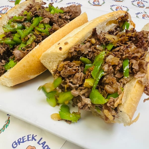 Two Greek Lady cheesesteak sandwiches, filled with meat, green peppers, and onions, on a white plate.