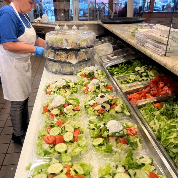 A Greek Lady employee prepares multiple plastic containers of fresh Greek salads with feta, olives, and eggs.