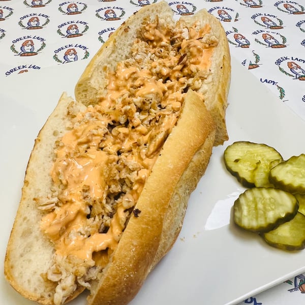A Greek Lady chicken sandwich with orange sauce and pickles on a white plate.