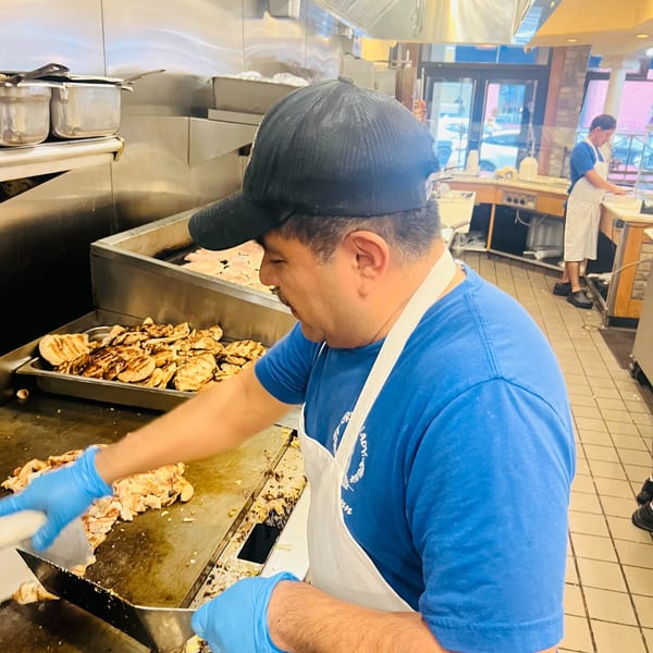 A Greek Lady chef in a blue shirt and apron cooks chicken on a large griddle.