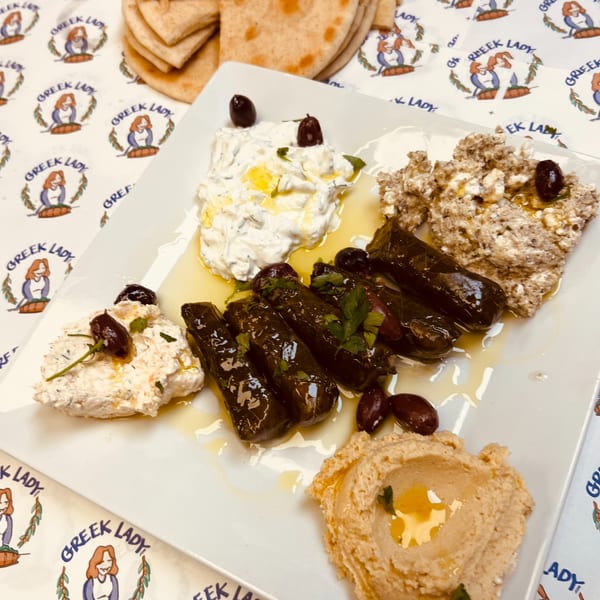 A Greek Lady appetizer platter with dolmades, hummus, tzatziki, and other dips, garnished with olives.